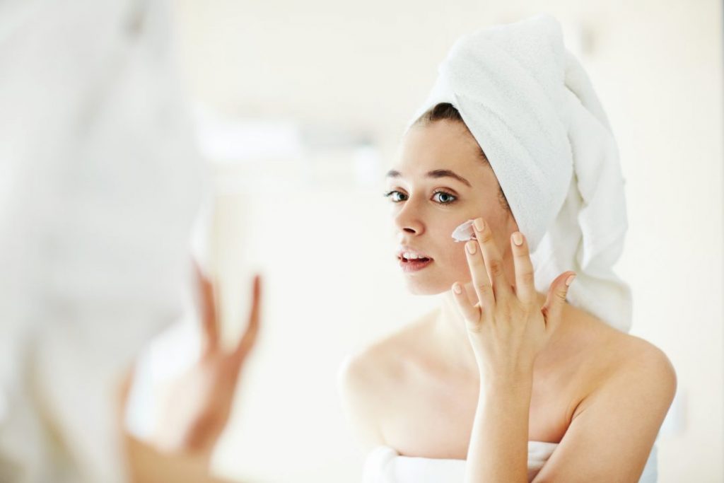 Mulher na frente do espelho passando creme facial | Como amenizar marcas de acne